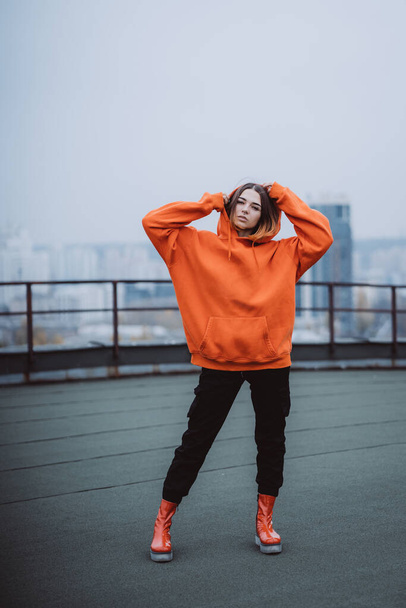 Dziewczyna w pomarańczowej kurtce pozuje na dachu budynku w centrum miasta - Zdjęcie, obraz