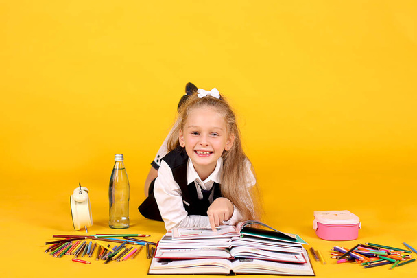 Siedmioletnia uczennica studiuje leżąc ze stosem otwartych książek na żółtym tle z kolorowymi ołówkami, piórnikiem, budzikiem i butelką wody. - Zdjęcie, obraz