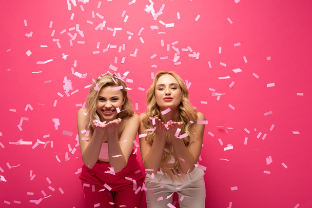 glimlachende blonde meisjes blazen lucht kus op camera onder vallen confetti op roze achtergrond - Foto, afbeelding