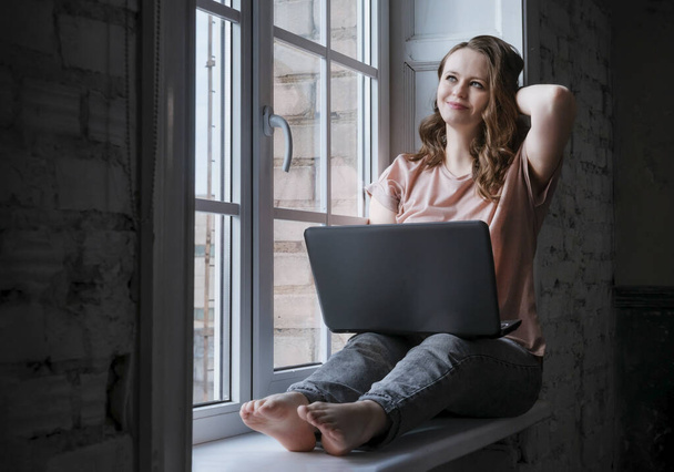1 white young woman in jeans with wavy hair sitting at the window with a laptop and smiling - Fotografie, Obrázek