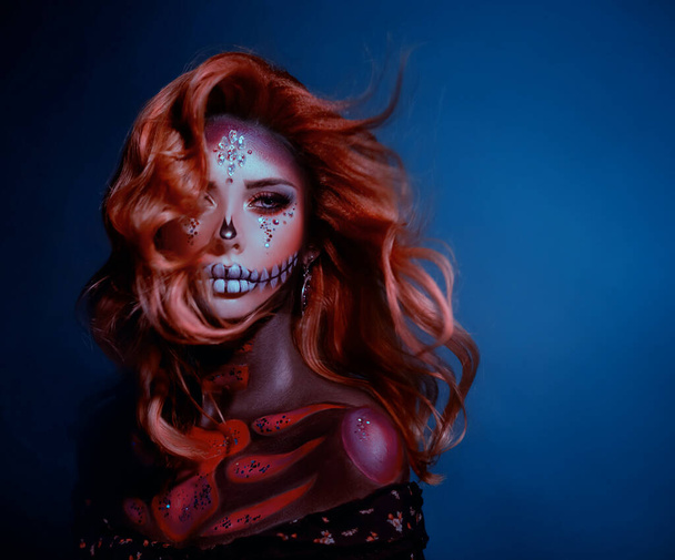 Artistic portrait young woman image sugar skull. Long redhaired wavy hair flies in wind. Stage creative makeup decorated with bright shiny sparkling crystal rhinestones. Backdrop blue color. Halloween - Foto, imagen