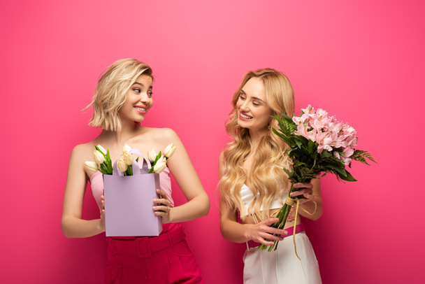 Blondýny dívky s kyticí usmívající se na sebe na růžovém pozadí - Fotografie, Obrázek