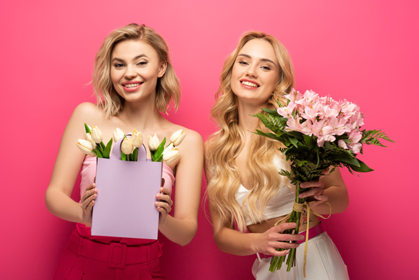 Beautiful blonde friends with bouquets smiling at camera on pink background - Foto, Imagem