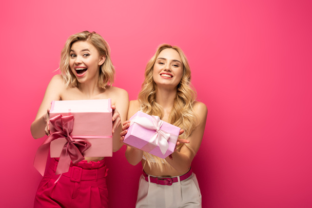 Filles blondes positives et choquées tenant des boîtes-cadeaux et regardant la caméra sur fond rose
 - Photo, image