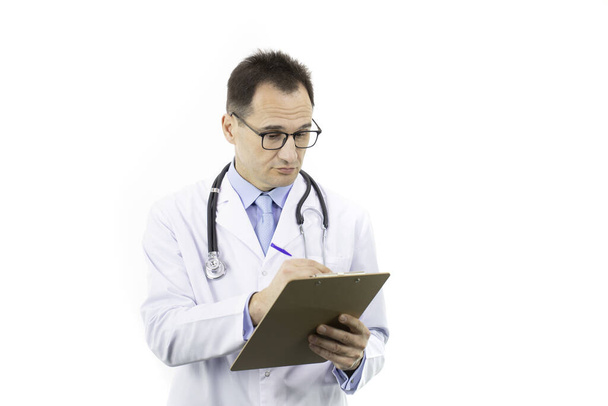Σοβαρός ιατρός με λευκό παλτό και στηθοσκόπιο που γράφει στο πρόχειρο - Φωτογραφία, εικόνα