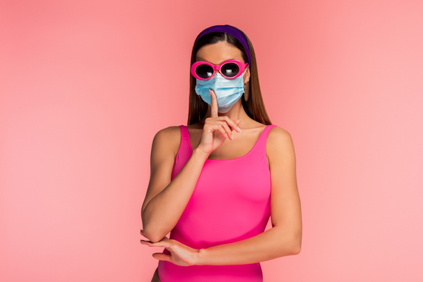 Vista frontal de la mujer en gafas de sol y máscara médica que muestra el signo shh aislado en rosa
 - Foto, imagen