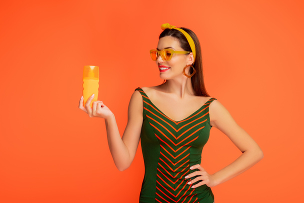 Widok z przodu kobiety z ręką na biodrze patrzącej na butelkę kremu do opalania i uśmiechającej się odizolowanej na pomarańczowo - Zdjęcie, obraz