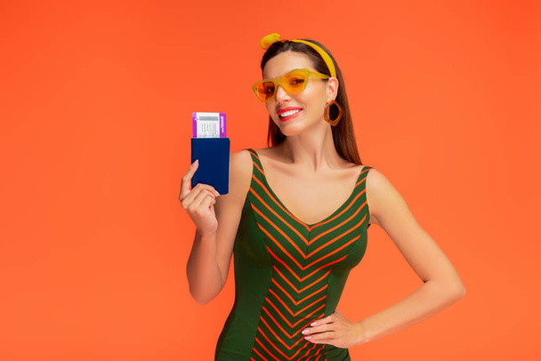 Widok z przodu kobiety z ręką na biodrze uśmiechnięta i pokazująca paszport z biletem lotniczym odizolowanym na pomarańczowo - Zdjęcie, obraz