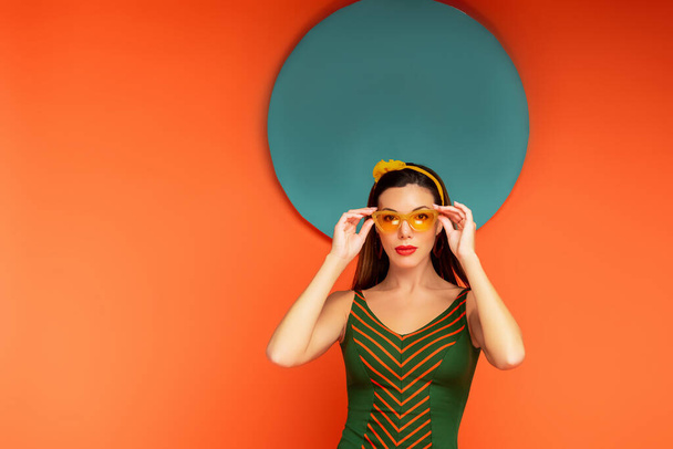 Woman touching sunglasses and looking at camera with blue circle behind on orange background - Valokuva, kuva
