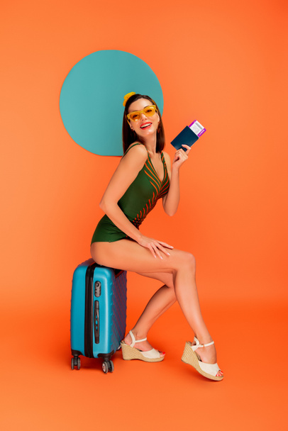A lány útlevéllel és repülőjeggyel mosolyog, a kamerába néz, és ül a bőröndön kék kör mögött a narancs  - Fotó, kép