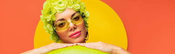 Portrait of girl in hat with decorative flowers and sunglasses looking at camera on yellow and orange background, panoramic shot - Photo, Image