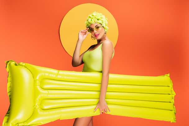 Girl in hat with decorative flowers smiling, touching sunglasses and holding inflatable mattress with yellow circle behind on orange - Valokuva, kuva
