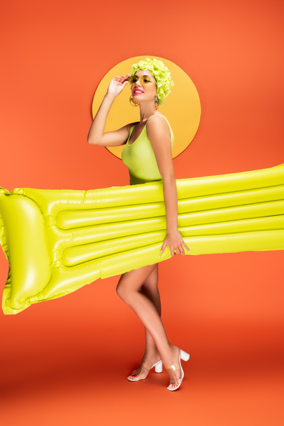 Girl touching sunglasses and holding inflatable mattress with yellow circle behind on orange - Foto, Imagen