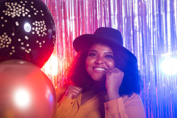 Jonge zwarte vrouw ziet er gelukkig uit terwijl ze in nachtclub zit. Verjaardagsfeest en nachtleven concept. - Foto, afbeelding