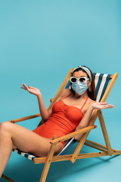 Confused woman in sunglasses and medical mask sitting on deckchair on blue background - Photo, Image