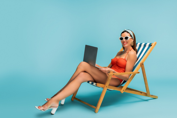 Freelancer with crossed legs and laptop smiling on deckchair on blue - Foto, immagini