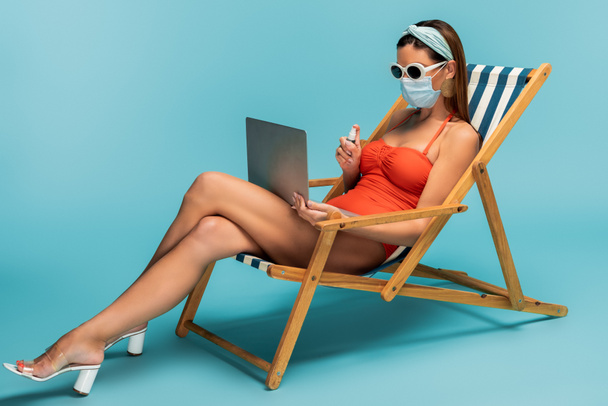 Freelancer in medical mask with laptop and bottle of hand sanitizer on deckchair on blue - Foto, afbeelding