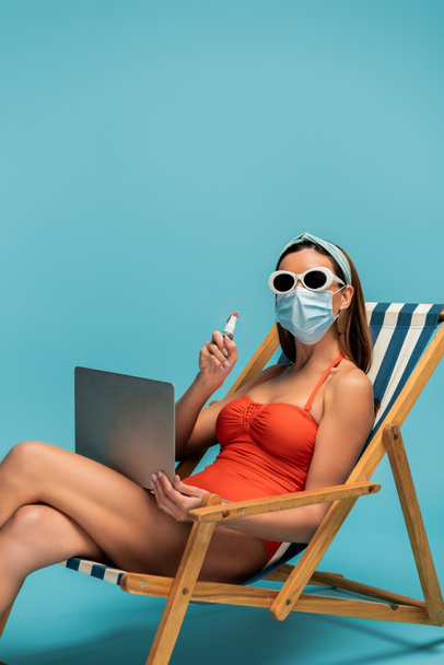 Freelancer in medical mask with laptop showing bottle of hand sanitizer on deckchair on blue - Foto, Imagem