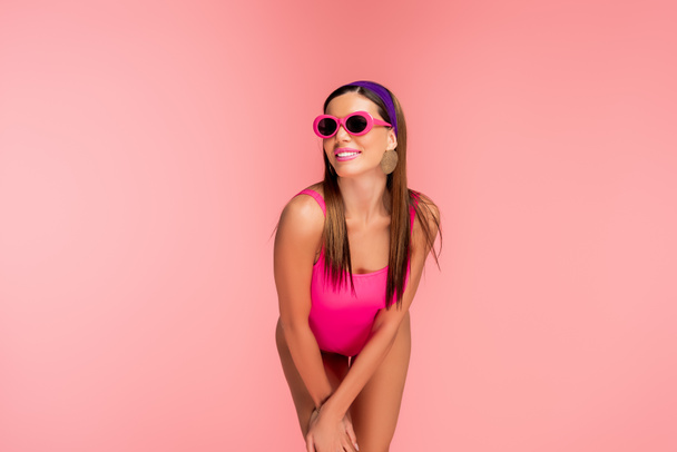 Женщина в солнцезащитных очках улыбается и наклоняется вперед изолированы на розовый
 - Фото, изображение