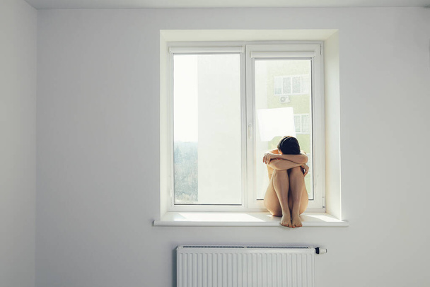 Θλιβερή γυναίκα αγκαλιάζει το γόνατό της και κλαίει στο παράθυρο - Φωτογραφία, εικόνα