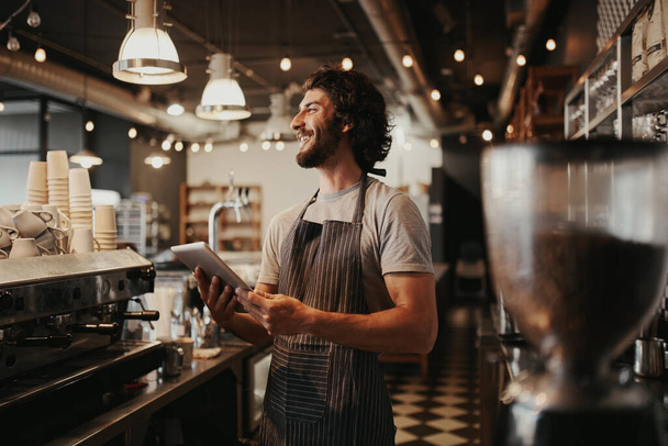 Handsome caucasian man in coffee shop laughing while holding digital tablet in hand standing behind counter - Foto, afbeelding