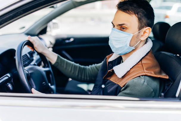 Beautiful young masked man sitting in a car, protective mask against coronavirus, driver on a city street during a coronavirus outbreak, covid-19 - Фото, изображение