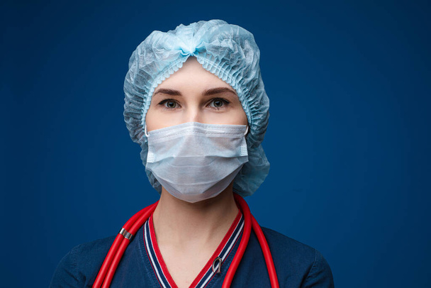 Beautiful woman doctor in white medical clothes, mask, phonendoscope and medical hat, picture isolated on blue background - Photo, Image