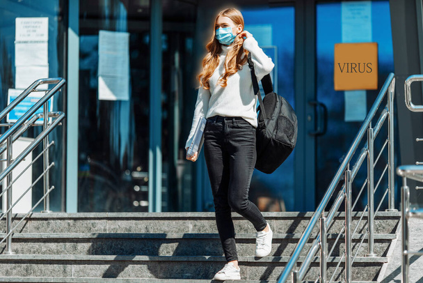 Beautiful young girl in a mask from coronovirus, student on the background of educational envy, women returning from work, remote work, covid-19, against the background of a glass business building - Zdjęcie, obraz