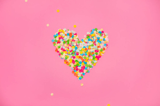Heart shape made of colorful confetti. Pink backdrop with scattered paper circles. Beautiful decor for the party. Festive backgrond for love story. Valentines day symbol. View from above. Flat lay. - Foto, afbeelding