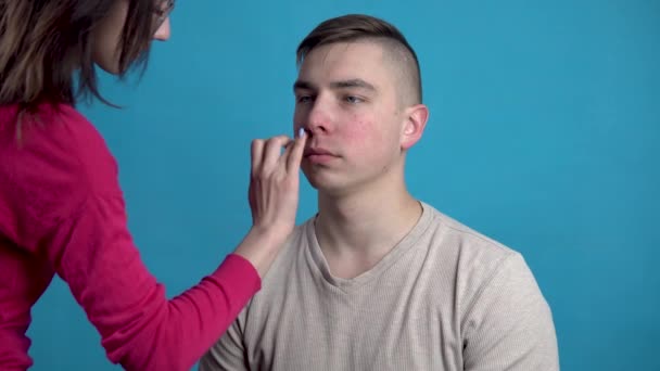 Make-up wordt toegepast op een jongeman. Het meisje past make-up op een man voor het schieten op een blauwe achtergrond. - Video