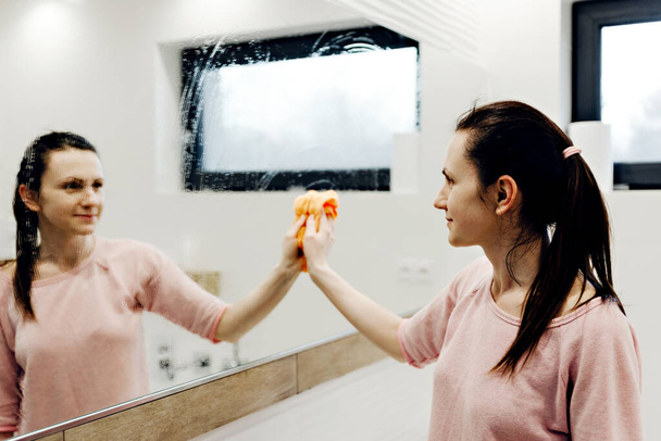 young housewife washes a mirror with detergent,happy woman cleaning mirror with rag at home,pretty woman cleans mirror in bathroom at home,cleaning the bathroom,mirror cleaning - Foto, Imagen