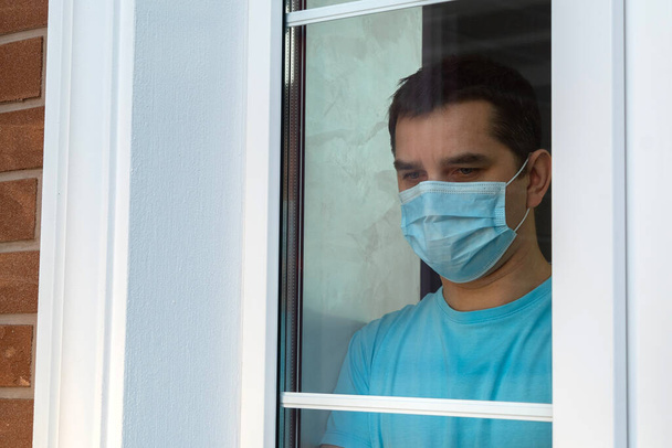 Κατάσταση έξαρσης του ιού του κερατοειδούς. Μολυσμένος άνθρωπος σε αυτο-απομόνωση κοιτάζει στο δρόμο μέσα από το παράθυρο του σπιτιού. Ένας άντρας με ιατρική μάσκα κοντά στο παράθυρο. Άντρας με χειρουργική μάσκα κοιτάζει έξω από το παράθυρο του σπιτιού του. - Φωτογραφία, εικόνα