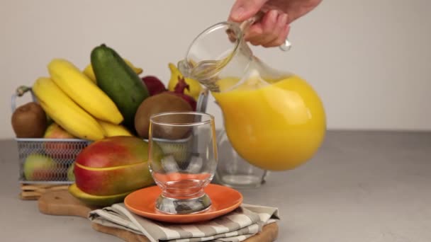 Pour juice from a decanter in glass. A glass of juice. Yellow refreshing summer drinks. Fresh Mango juice, orange juice. Fruit basket on the table. Fresh fruits and juices in the kitchen. Vegan Food. Vitamin drink. - Filmati, video
