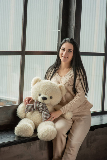 Young attractive woman posing in fashionable clothes near the window. Girl at the window in a beige knitted suit. Brunette, beautiful young girl, portrait. Girl with a soft plush toy bear in her hands - Фото, изображение