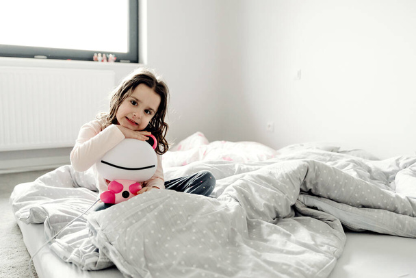 young girl woke up and lies in bed with a toy,young girl lying on the bed,child hugs a toy,joyful girl smiling and sitting on the bed,weekend morning - Photo, Image
