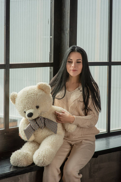 Young attractive woman posing in fashionable clothes near the window. Girl at the window in a beige knitted suit. Brunette, beautiful young girl, portrait. Girl with a soft plush toy bear in her hands - Fotoğraf, Görsel