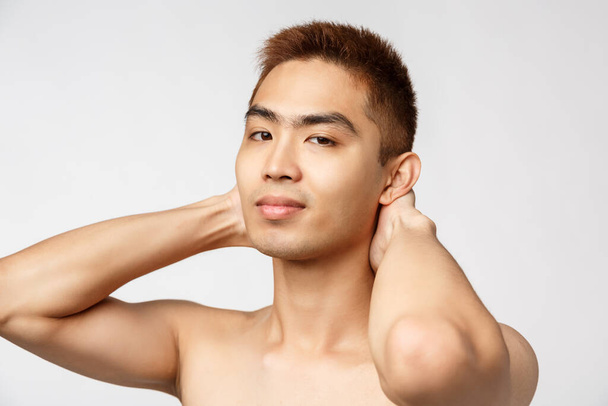 Beauty, skincare and men health concept. Portrait of handsome young asian man standing naked torso, hold hands behind head and smiling camera pleased, looking after appearance - Foto, afbeelding