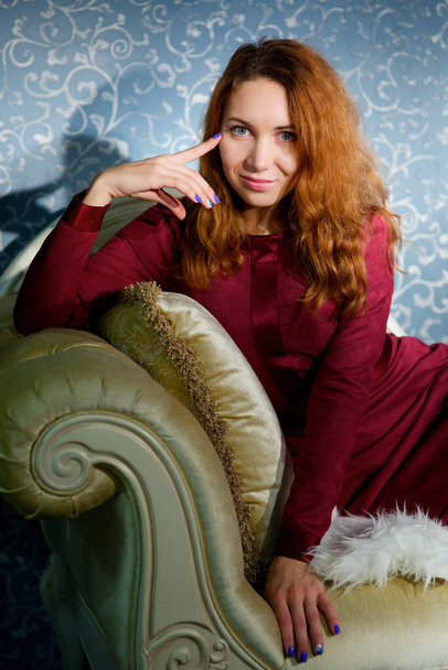 Una chica con un vestido rojo se sienta en un viejo sofá vintage en la habitación
. - Foto, Imagen