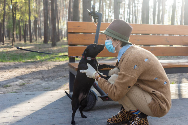Eine stilvolle Frau mit Maske und Handschuhen, um sie vor dem Coronavirus zu schützen. Mit Elektro-Roller in der Stadt und Park, um sicher zu sein. Spaß mit ihrem Hund. - Foto, Bild