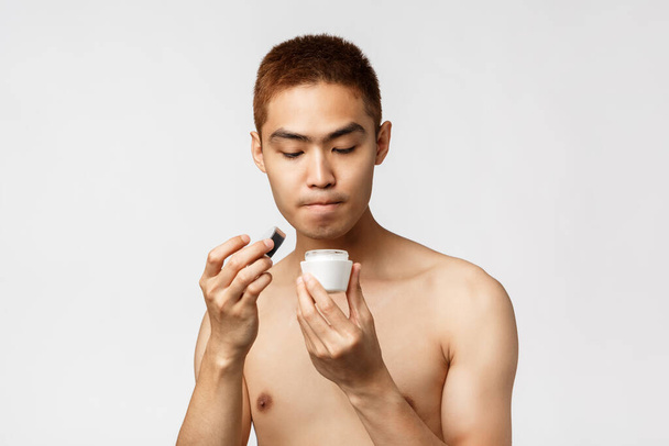 Krása, lidé a volnočasové pojetí. Portrét zvědavého asijského muže s nahým trupem otevřené přítelkyně krém na obličej, použít kosmetický přípravek pro péči o pleť rutiny, stojící bílé pozadí - Fotografie, Obrázek