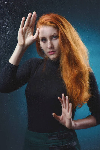 A beautiful red-haired girl in a black turtleneck is sad behind a glass with water drops, a photo of a glamorous young, slender woman behind a blind glass - Fotó, kép