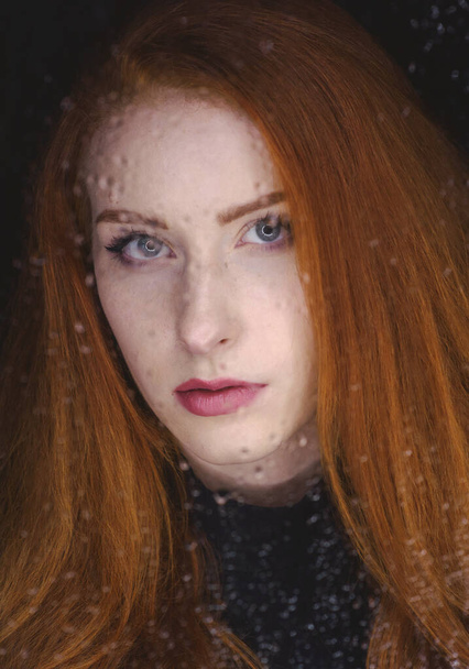 A beautiful red-haired girl in a black turtleneck is sad behind a glass with water drops, a photo of a glamorous young, slender woman behind a blind glass - Foto, afbeelding