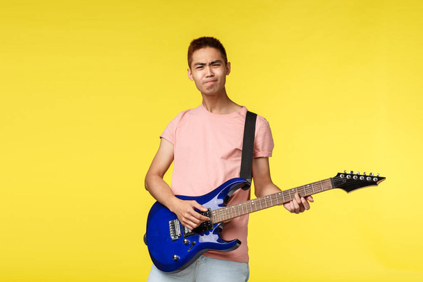 Lifestyle, leisure and youth concept. Enthusiastic carefree asian man in pink t-shirt playing on electric guitar over yellow background, enjoying performing with band, learn new instruments - Photo, Image