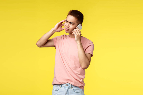 Tecnología, comunicación y concepto de estilo de vida. problemático asiático hombre en rosa camiseta, sentirse frustrado, hablar en el teléfono móvil, entrecerrar los ojos y muecas molestado, de pie indeciso amarillo fondo
 - Foto, Imagen
