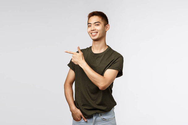 Pyšný dobře vypadající asijský student ukazující prstem vlevo a usmívající se fotoaparát potěšen, chlap ukazující cestu k uložení, on-line odkaz nebo firemní banner, představit položku, šedé pozadí - Fotografie, Obrázek