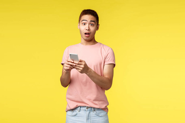 Technologia, komunikacja i koncepcja stylu życia. Zaskoczony azjatycki mężczyzna w różowym t-shirt, trzymając telefon komórkowy, otwarte usta dysząc zastanawiał, reagować na ciekawe wiadomości online, żółte tło - Zdjęcie, obraz
