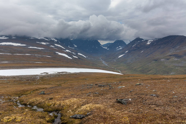 Beautiful wild nature of Sarek national park in Sweden Lapland with snow capped mountain peaks, river and lake, birch and spruce tree forest. Early autumn colors in stormy weather. selective focus - 写真・画像