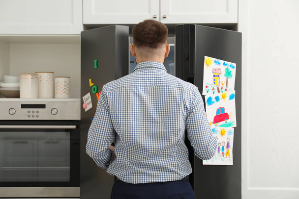 Mann öffnet Kühlschranktür mit Kinderzeichnungen, Notizen und Magneten in Küche - Foto, Bild