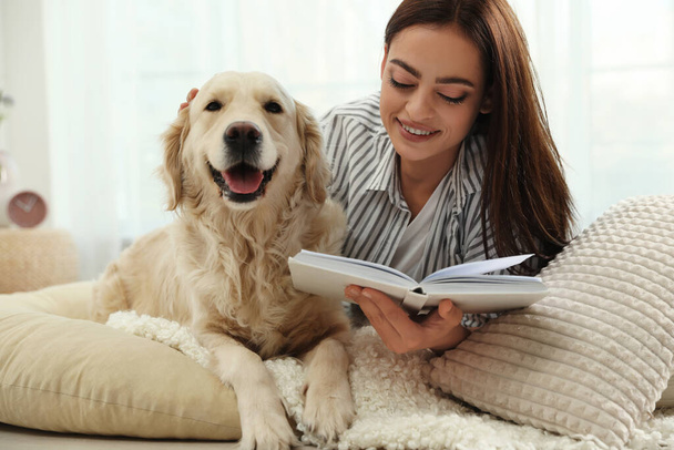 Young woman with book and her Golden Retriever at home. Adorable pet - Zdjęcie, obraz