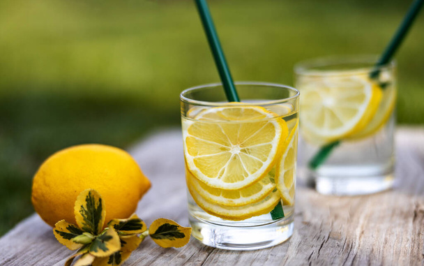 夏の飲み物、レモンのスライスでいっぱいの水のグラス、晴れた暖かい日に芝生の上の木の板の上に立つと、背景がぼやけている、フィールドの浅い深さ、選択的な焦点. - 写真・画像